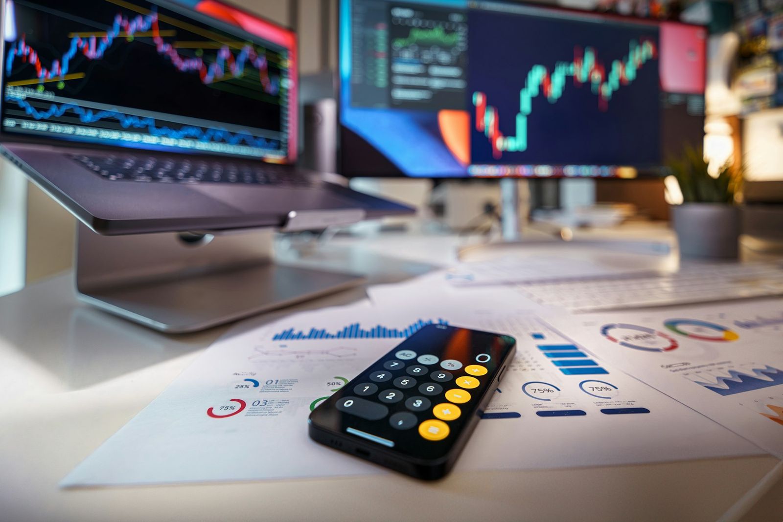 Calculator on printed financial reports, with market charts on screens behind